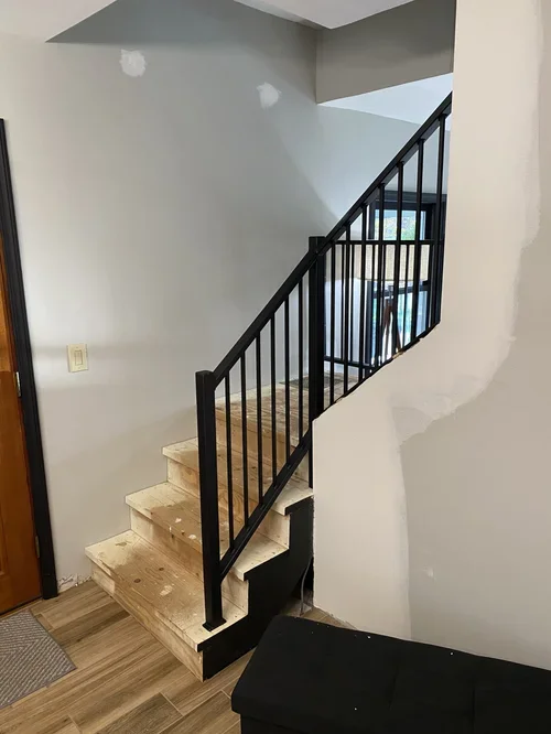 Interior view of a partially constructed staircase with wooden steps and a black metal railing in a house under renovation. There is an unfinished patch on the wall near the stairs and a black piece of furniture at the bottom.