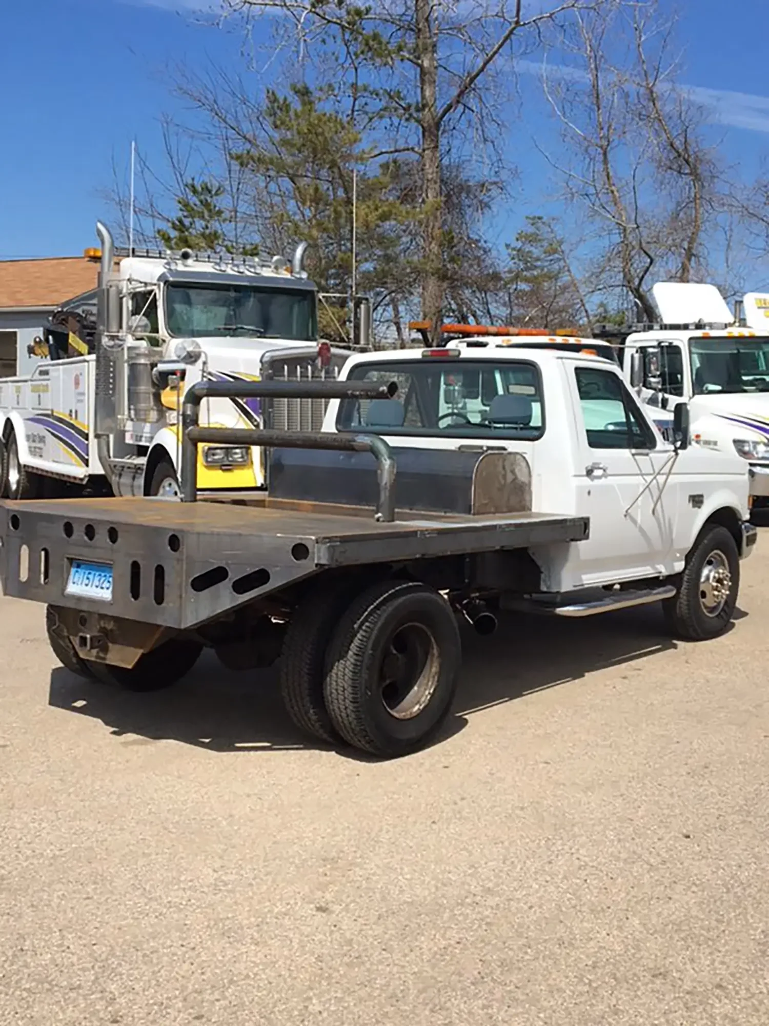 A white flatbed utility truck parked on a gravel lot with other utility trucks nearby, a tree line in the background, and a clear blue sky.