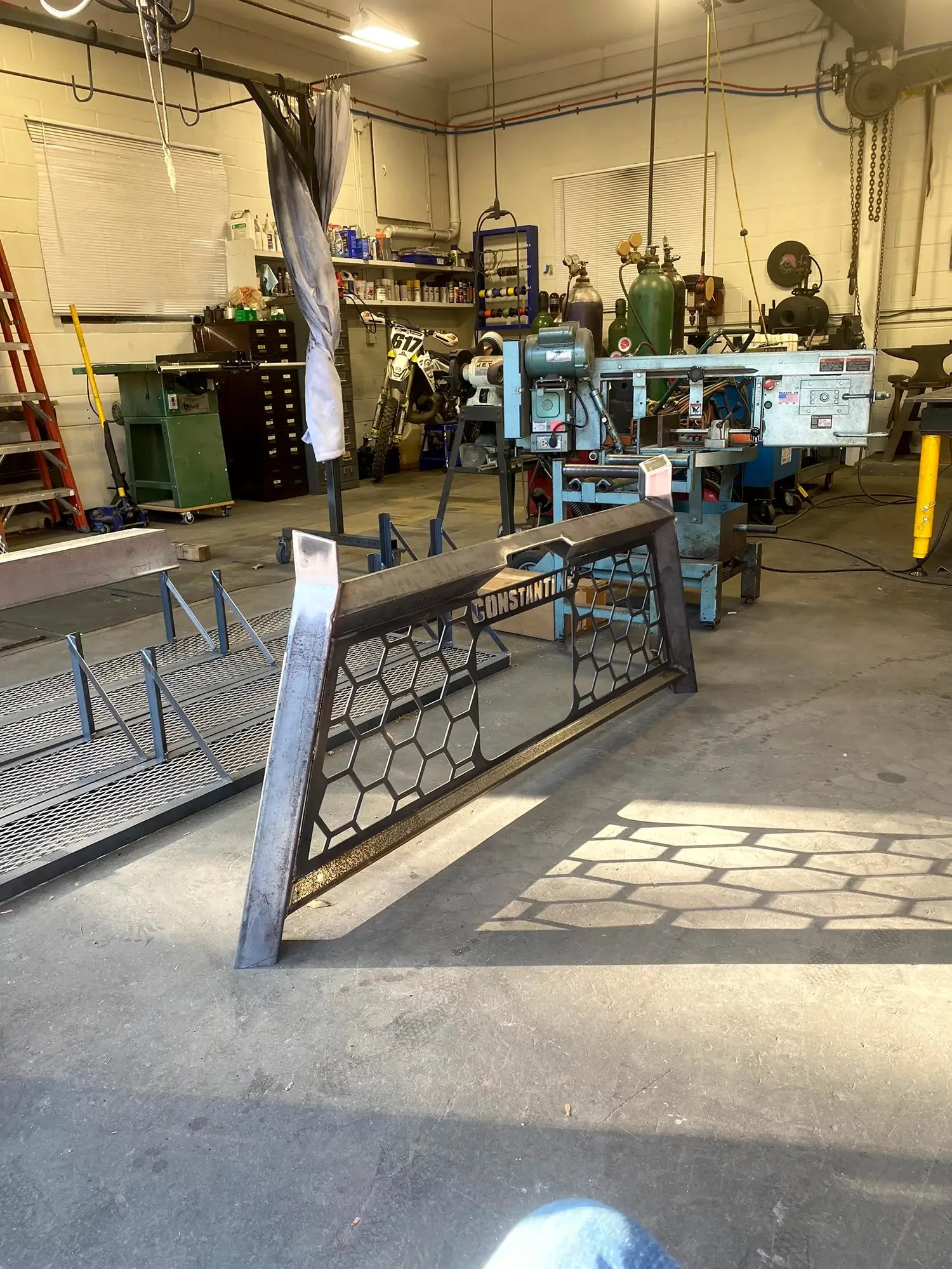 A workshop with various tools, parts, and equipment including a motorcycle, metal fencing, and a workbench with gas cylinders, a ladder, and storage shelves.