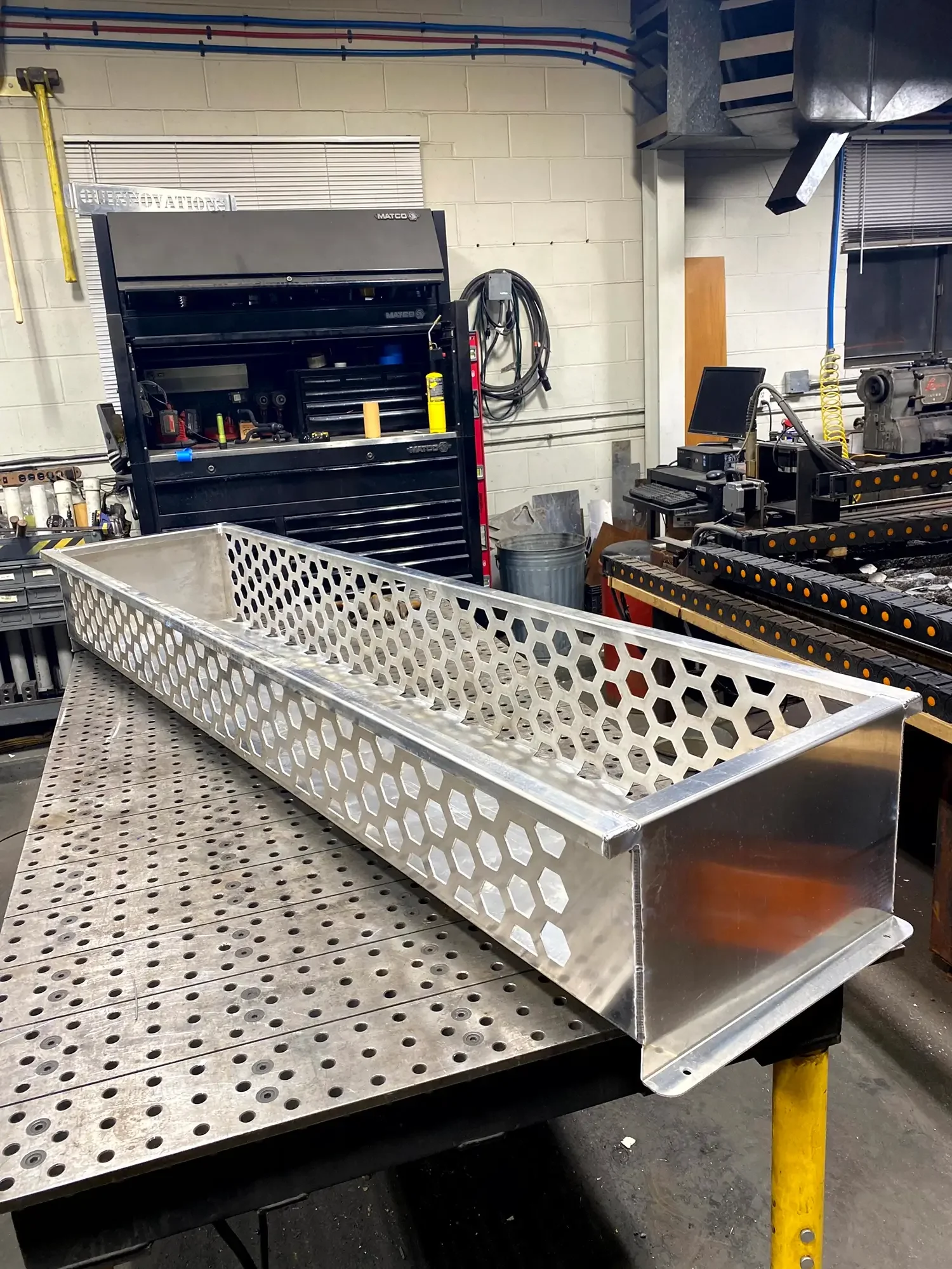 An empty silver metal tray with hexagonal cutouts on a perforated workbench in a workshop. In the background are tools, a computer, and industrial equipment.