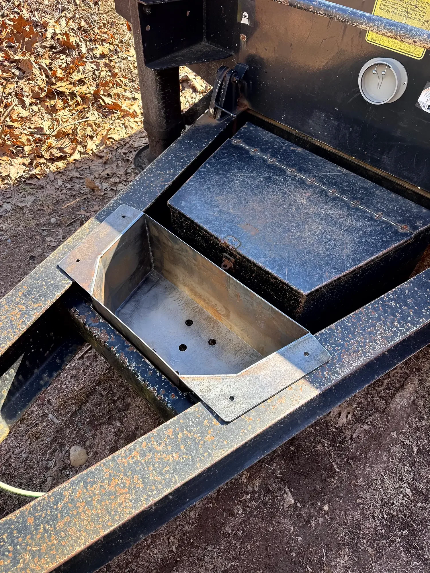 A black metal trailer with a built-in propane grill, situated on dirt ground. The grill has a silver metallic tray and a black lid.