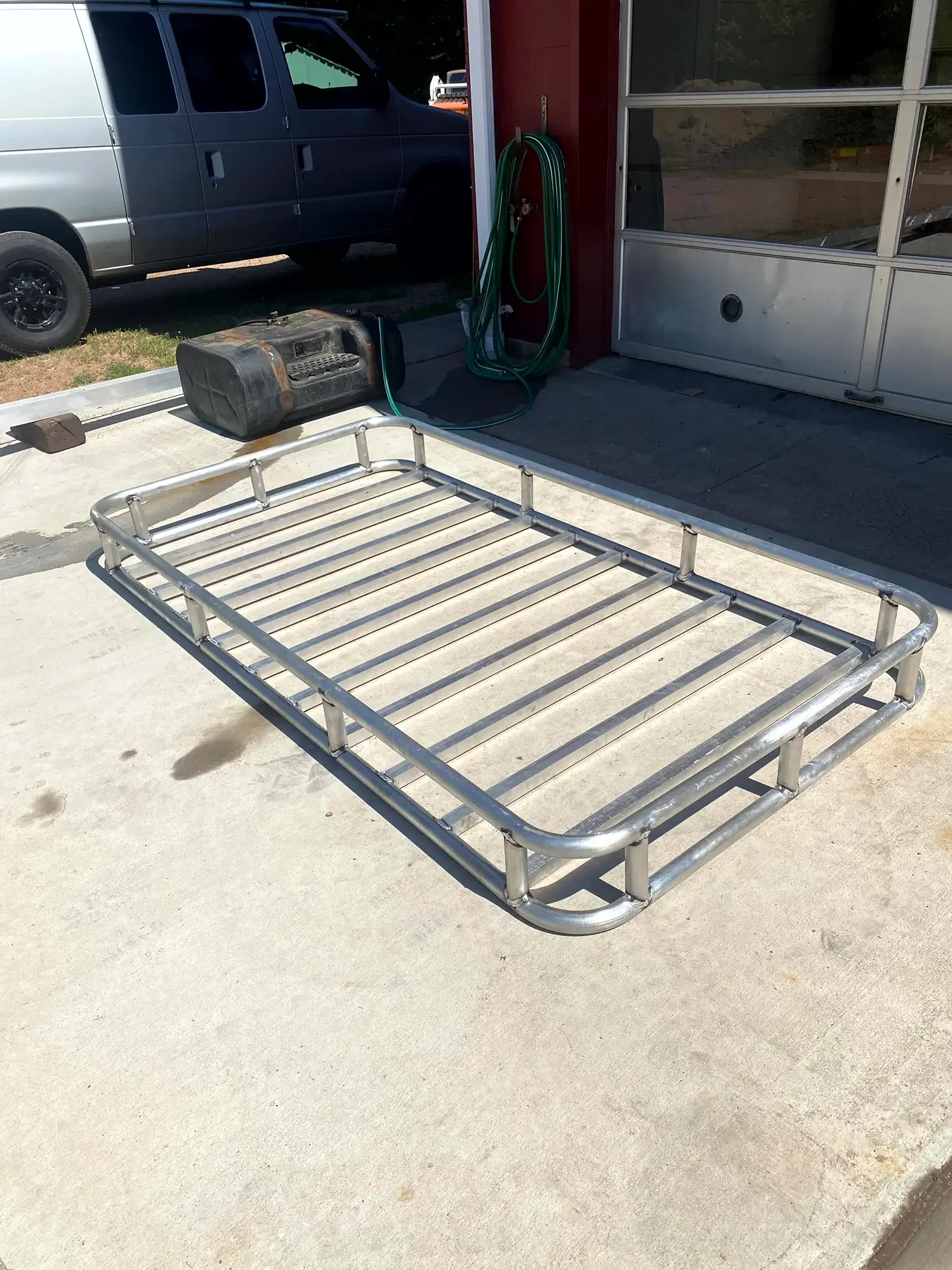 Empty metal bed frame on concrete ground, garage with tools and a black tank in the background, parked gray van outside with another vehicle partially visible.