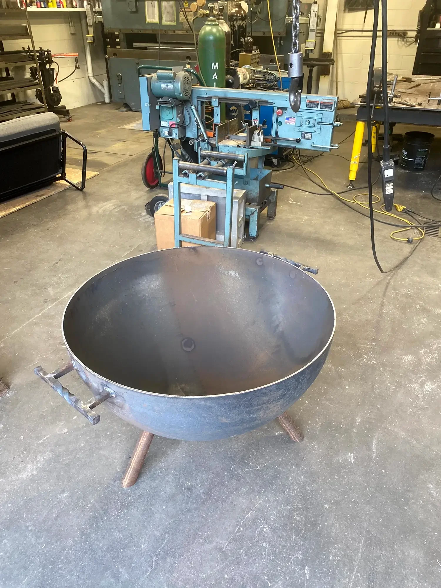 Empty large metal basin on a stand in a workshop or industrial space, with machinery and tools in the background.