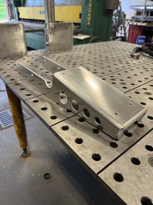 A metallic, anodized aluminum bracket or mount on a workbench in a workshop. The bracket has multiple holes and a curved shape, possibly for mechanical or structural purposes.