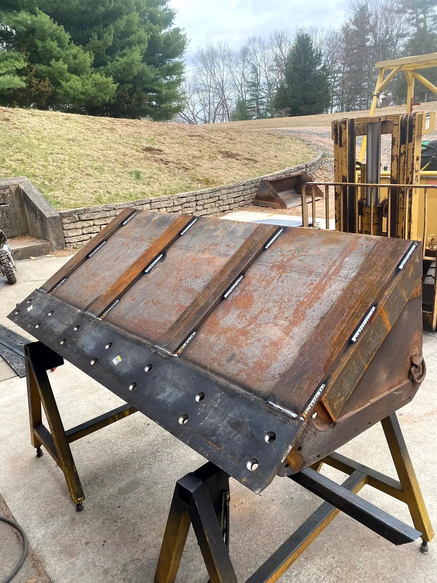 A metal workbench with a rusty surface, supported by a wooden sawhorse, with a large construction excavator in the background, outdoors on concrete with grassy hills and trees nearby.