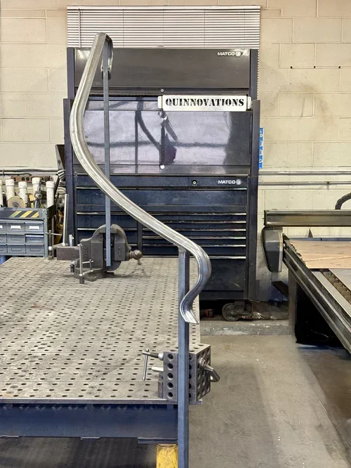 Workshop with metal work table, a large black tool chest with a sign labeled 'QUINNOVATIONS,' and a metal pipe bent in a spiral shape attached to the table.