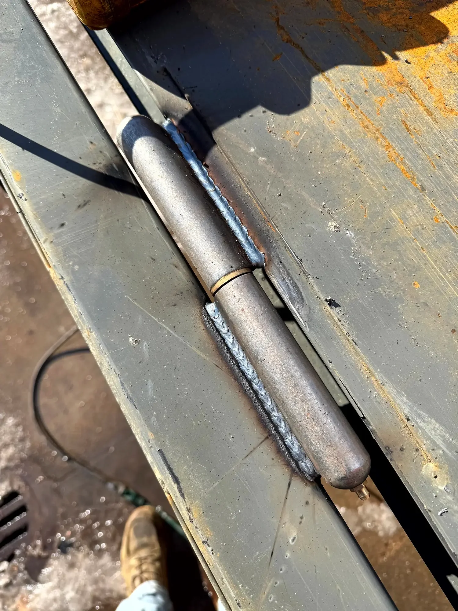 Close-up of a metal welded joint on a cylindrical metal pipe, with rust and scratches on the surface, on a gray metal surface, foot in the lower left corner, standing on a dirt ground.