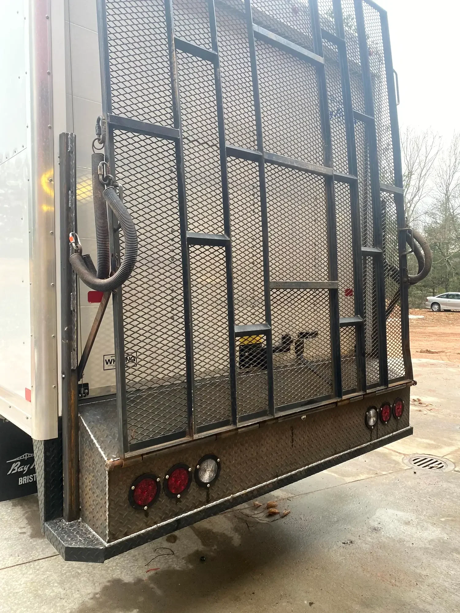 A black metal dog crate inside a trailer, with a dog lying down at the bottom, and some trees and a parked car visible outside in the background.