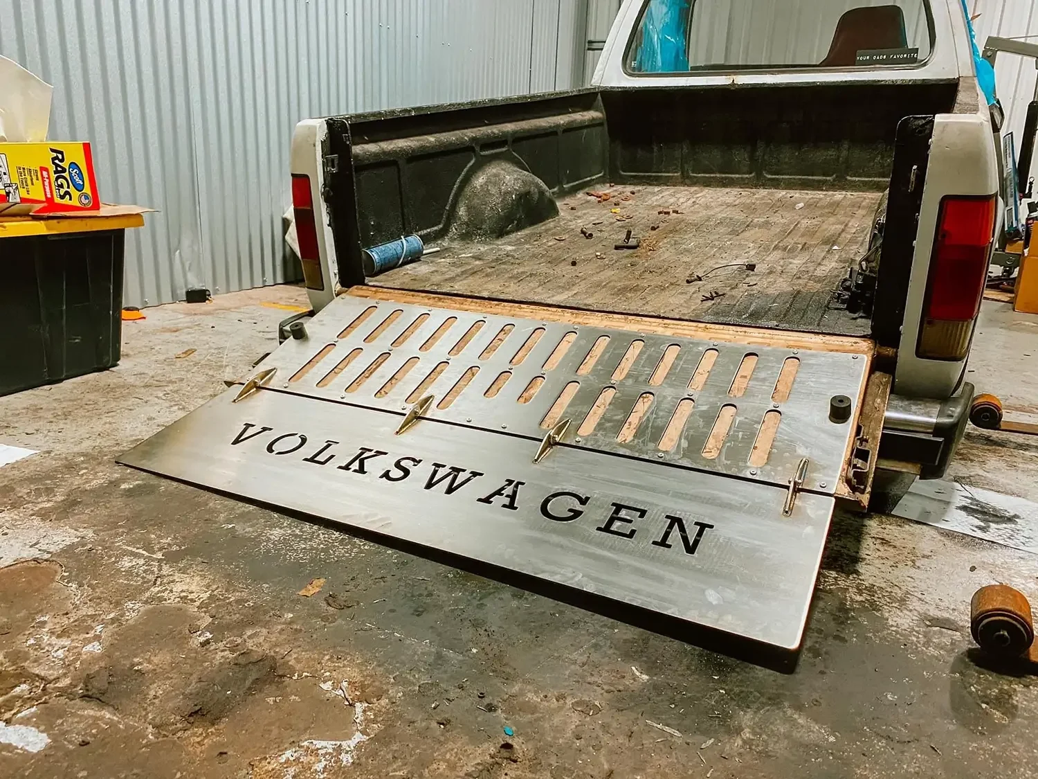 A pickup truck bed with a customized metal tailgate that reads 'Volkswagen,' placed on the ground inside a workshop. The tailgate has a slatted design with brass hardware.
