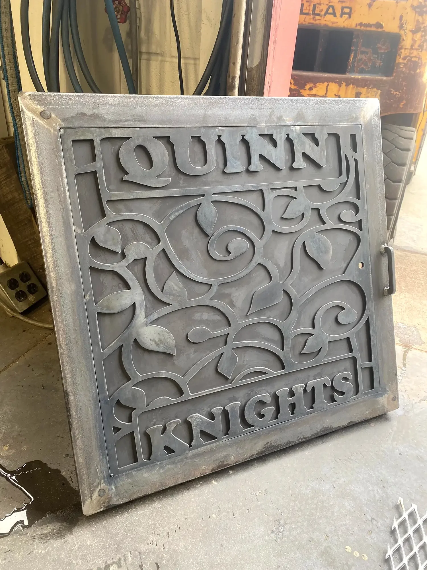 A metal decorative panel with the word 'HOUIN' and a partially visible word 'LIGHTS' at the bottom. The panel has intricate floral and swirl cutout designs. It leans against a wall or structure in a workshop or industrial setting.