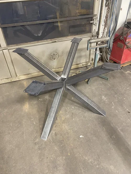 A metal structure resembling the base of a table or chair, with multiple angled legs and a rectangular platform on one side, placed on a workshop floor.