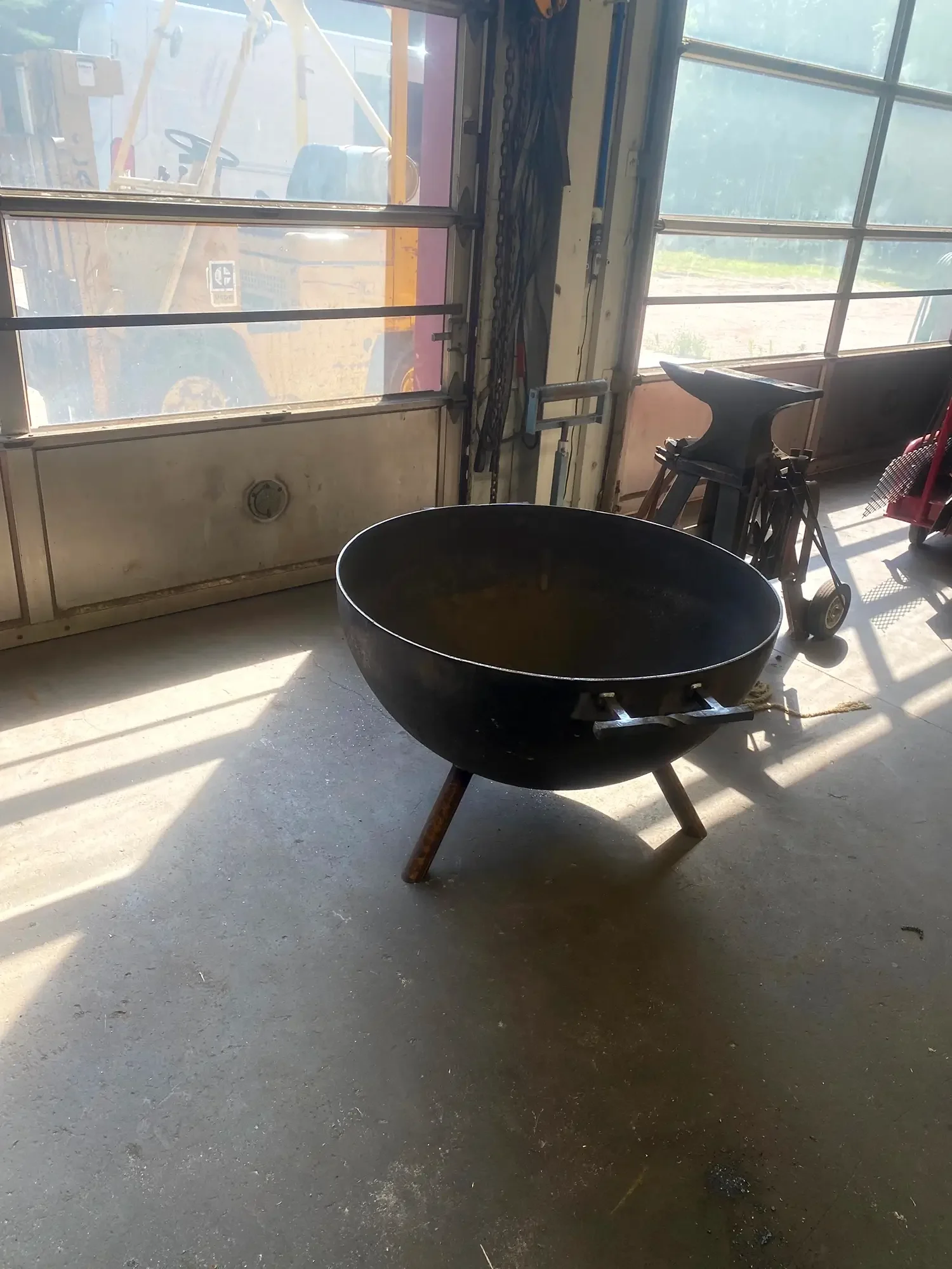 Black fire pit in a garage with sunlight coming through large windows, with an anvil, chains, and other tools nearby.