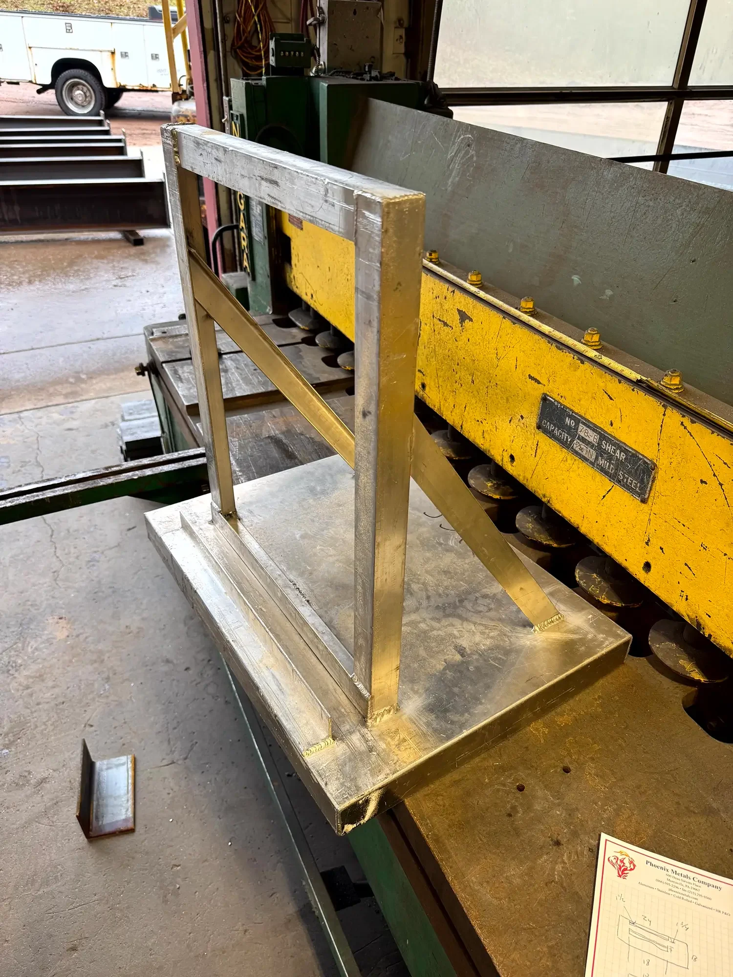 A metal frame positioned on a workbench in a workshop. In the background, there are various tools and machinery, including a yellow metal piece with bolts, a silver metal sheet, and a handle. Outside the workshop, a white utility truck is visible.
