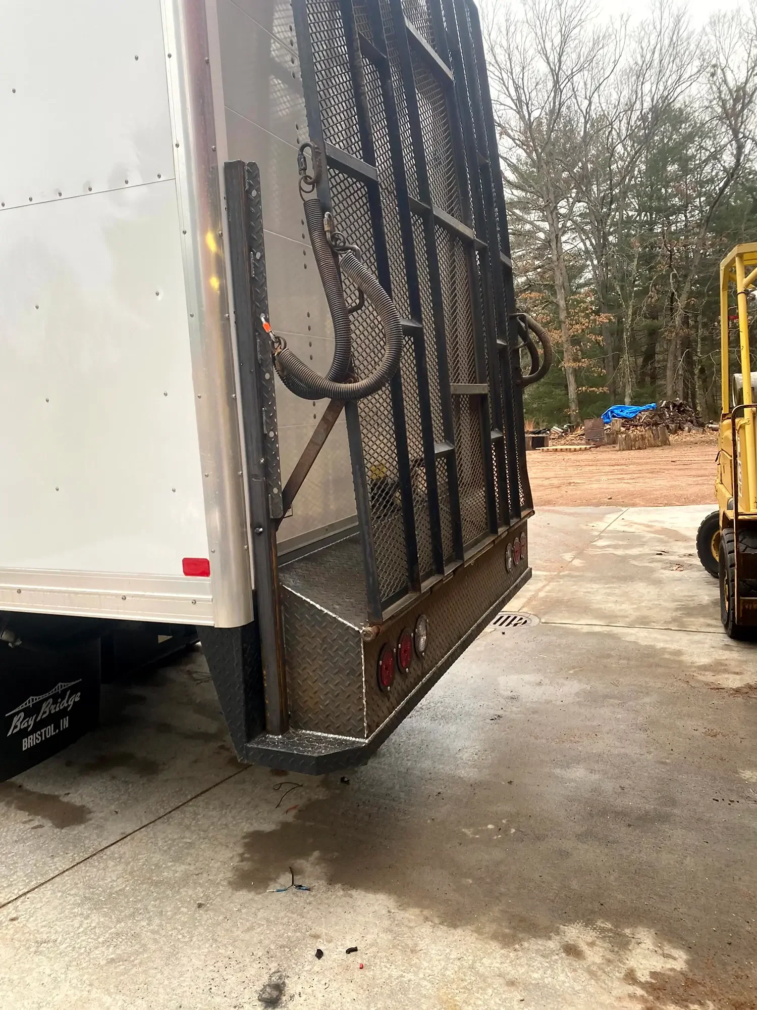 The side of a utility truck with black metal rack and hoses, parked outdoors with trees and dirt in the background.