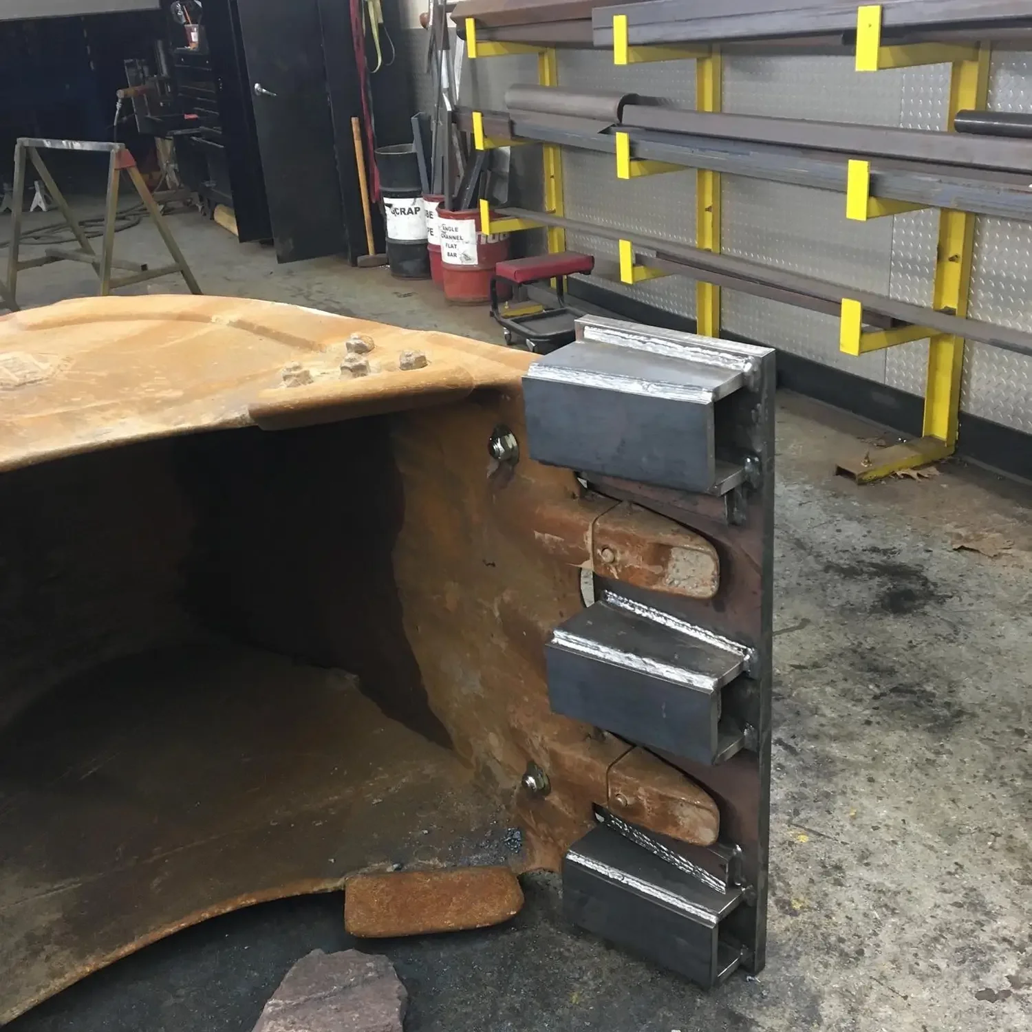 Industrial workspace with a large, rusted metal object and a metal support structure with rollers. Workshop tools and supplies are visible in the background.