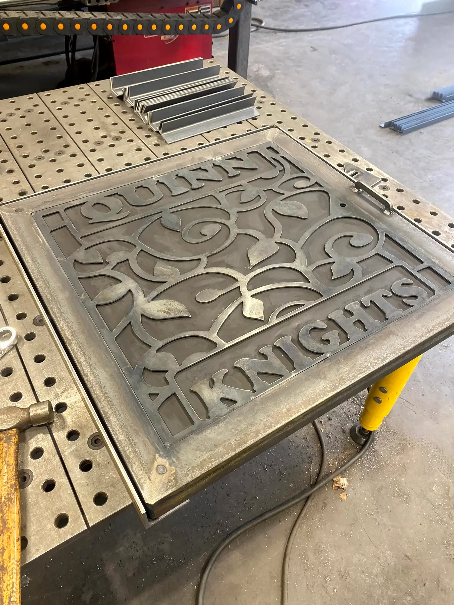 A metal worktable with a decorative metal cutout piece, several small metal L-shaped brackets, and a black and yellow adjustable clamp. The table has a pegboard surface with multiple holes and is situated on a concrete floor with some metal pieces ne