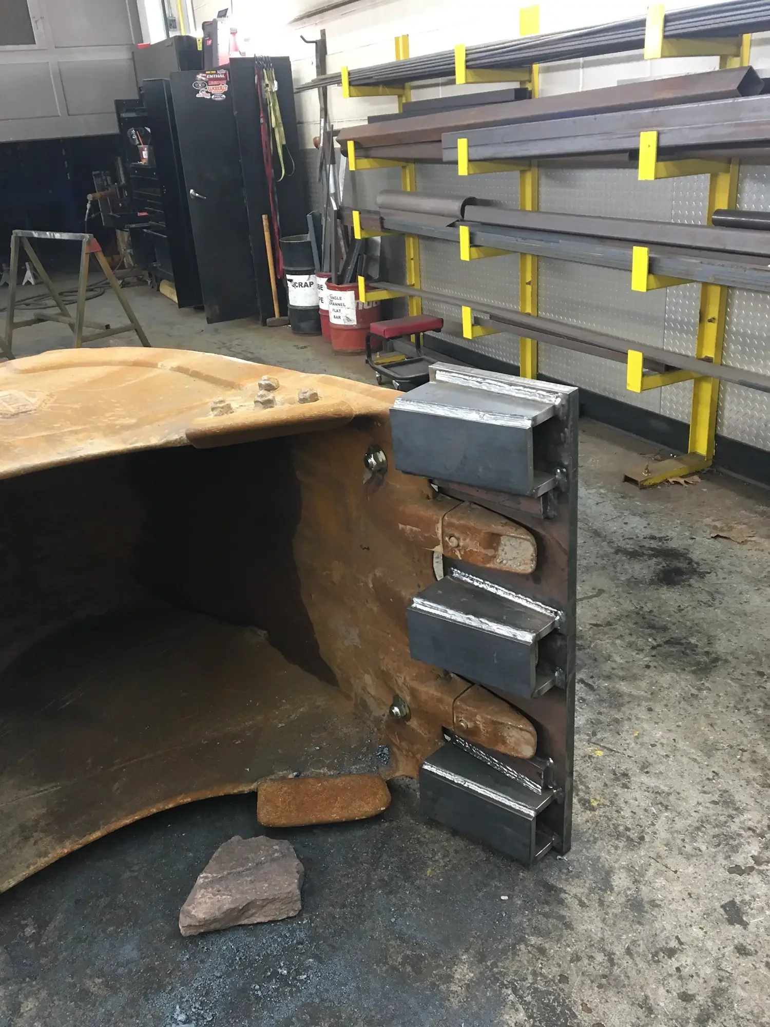 A workshop scene with a large wooden workpiece and metal clamps, yellow shelving with metal bars, black storage cabinet, and various tools and supplies.