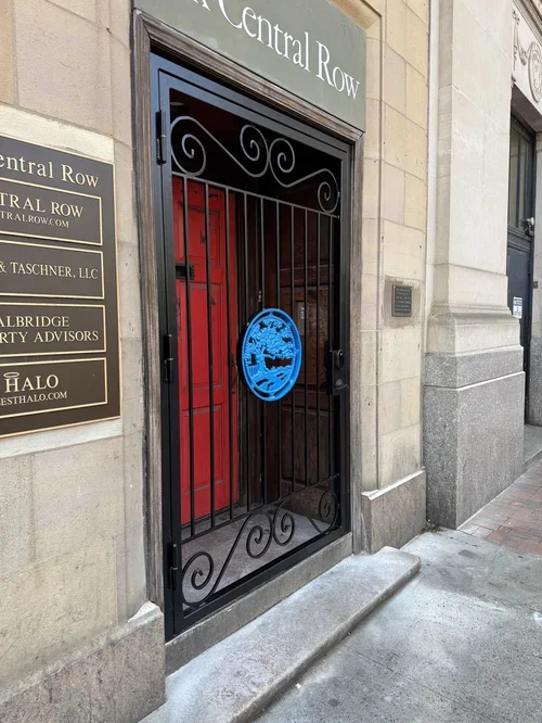 Black ornate security gate with the Central Row logo in blue, blocking the entrance to a building with signboards on the wall.