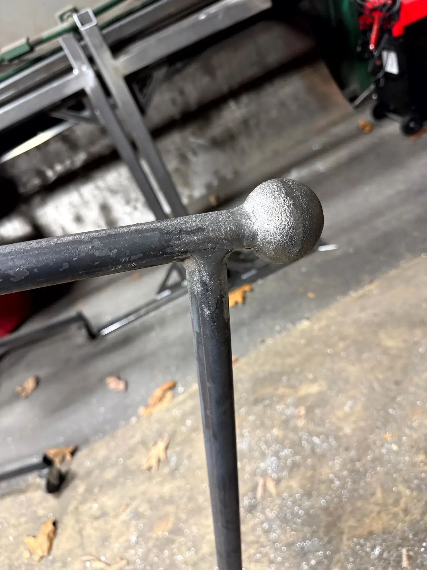 Close-up view of a black metal axe or pickaxe with a round ball-shaped end, resting against a metal shelving unit in a garage or workshop with a concrete floor and some fallen leaves.