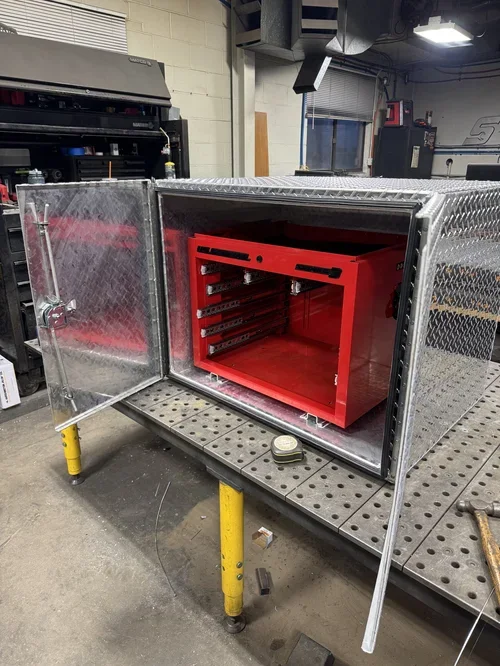 Metal enclosure with red interior, open door with chain latch, placed in an industrial workshop on a workbench.
