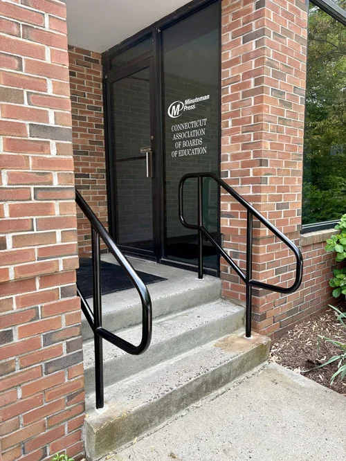 Entrance door to the Connecticut Association of Boards of Education office with black handrails on concrete steps and brick walls surrounding the door.
