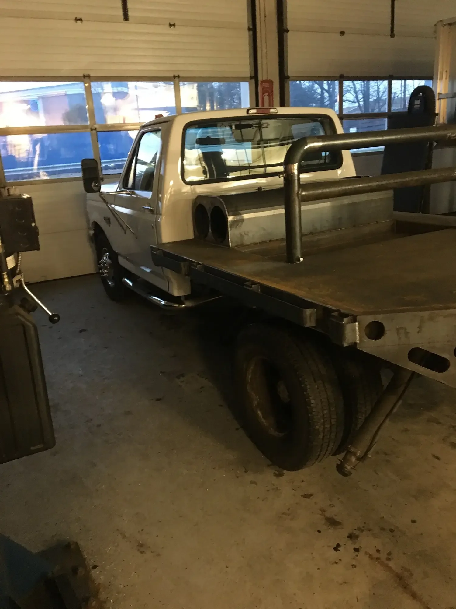 A small white pickup truck parked inside a garage with large windows, with a flatbed at the back and black tires.