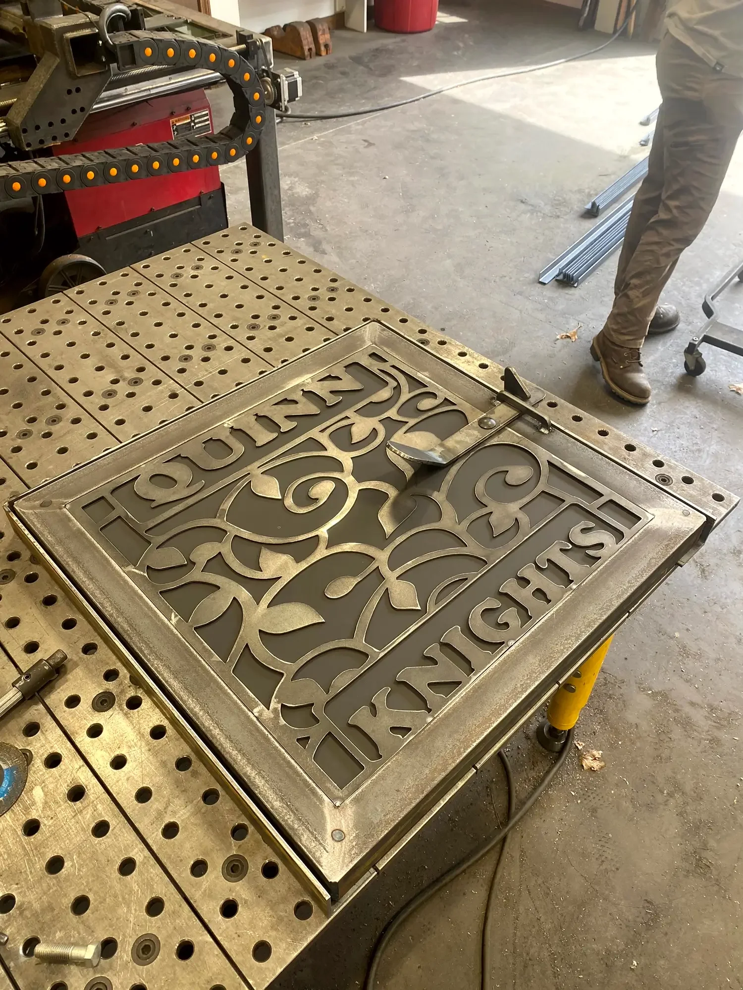 A metal table with a decorative metal panel and a welding or cutting tool on it, in a workshop setting. The panel has intricate scrollwork and the table has a perforated surface with holes. There are tools, a person, and other workshop items visible 