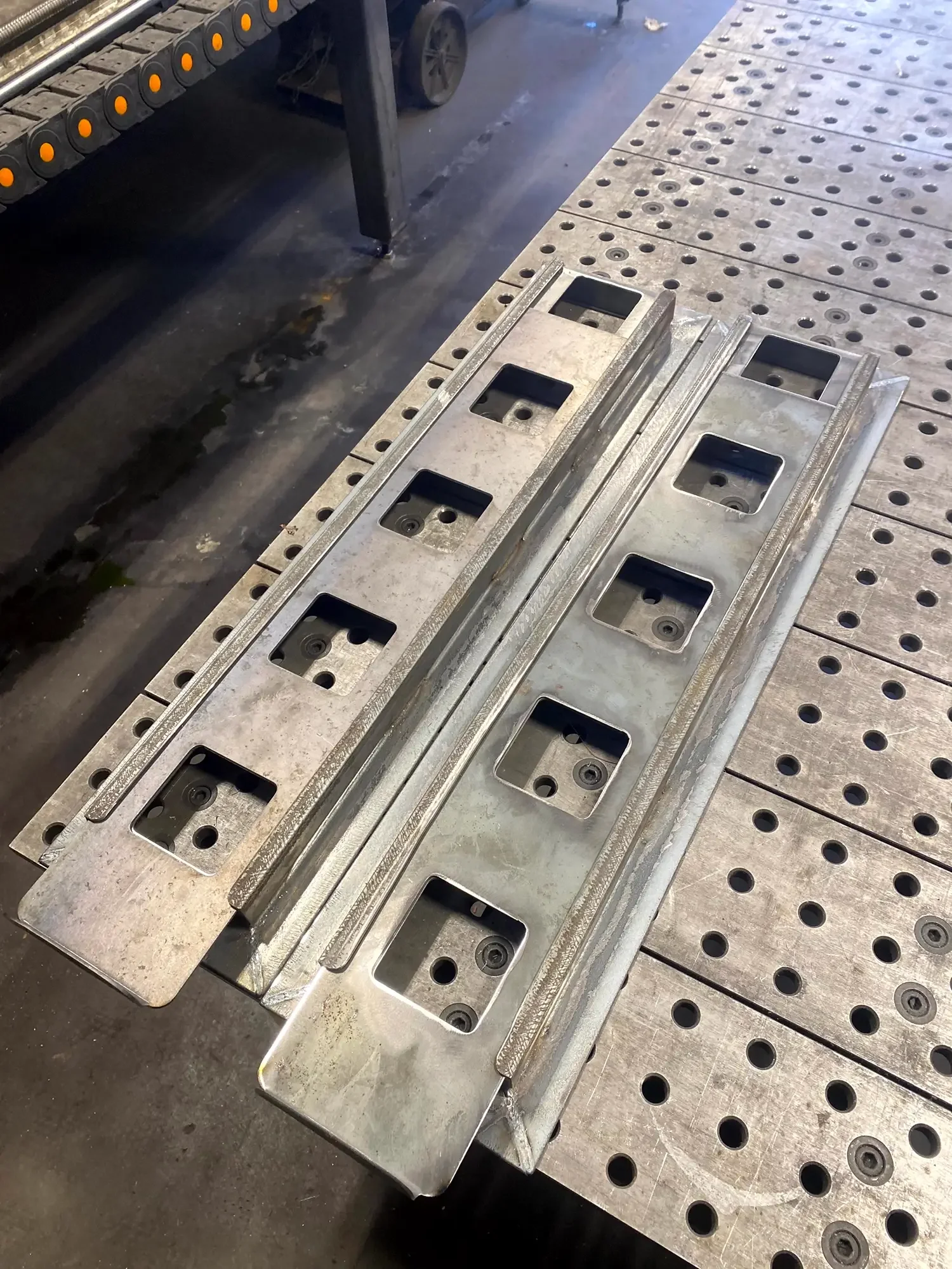 Metal parts with rectangular cutouts and holes, arranged on a workbench in an industrial setting.