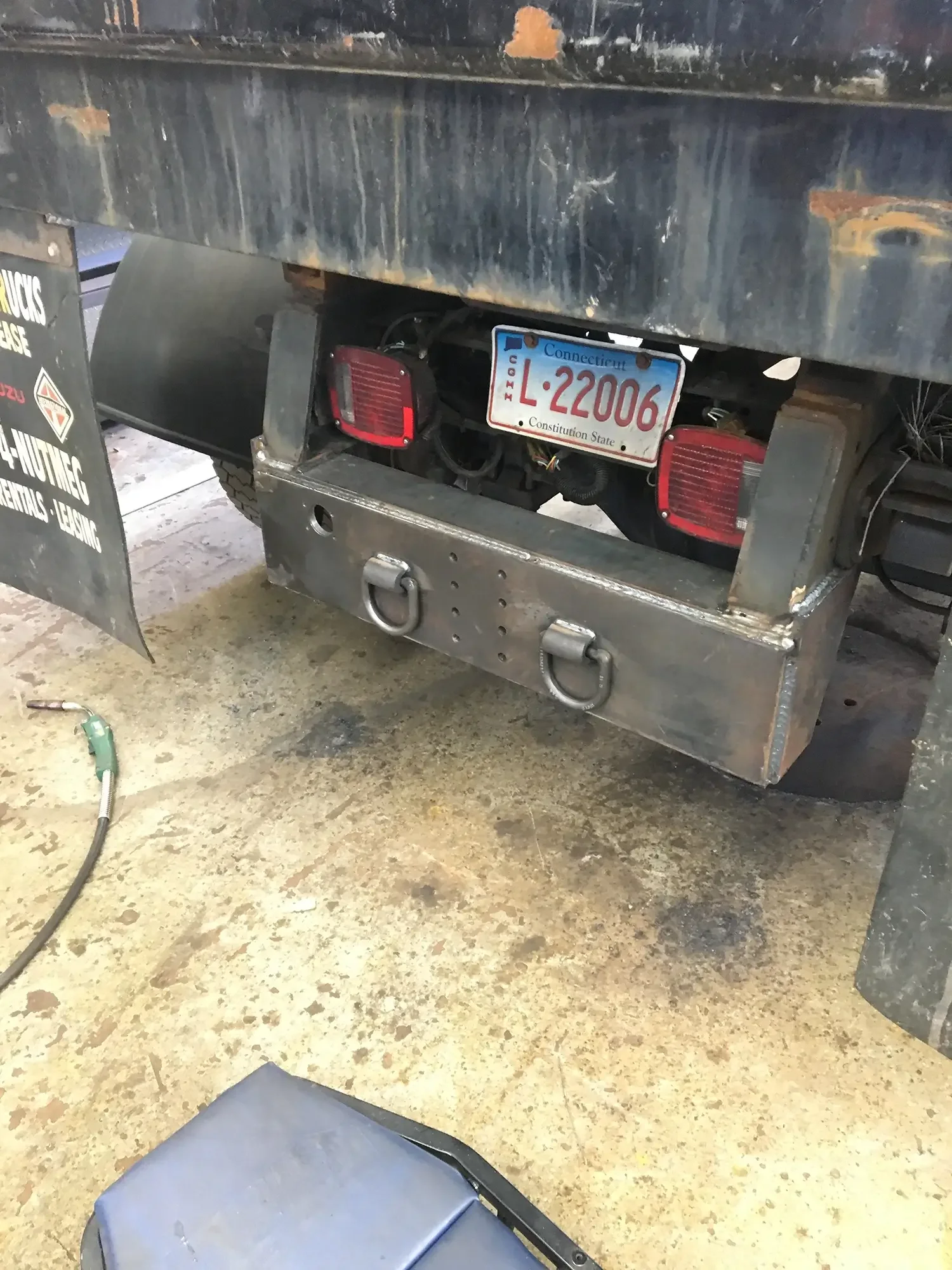 A metal trailer with a Connecticut license plate (L-22006) and red taillights, parked in a workshop. The trailer has a large, weathered black metal surface and a trailer hitch hook on the front. The workshop floor appears stained and slightly texture