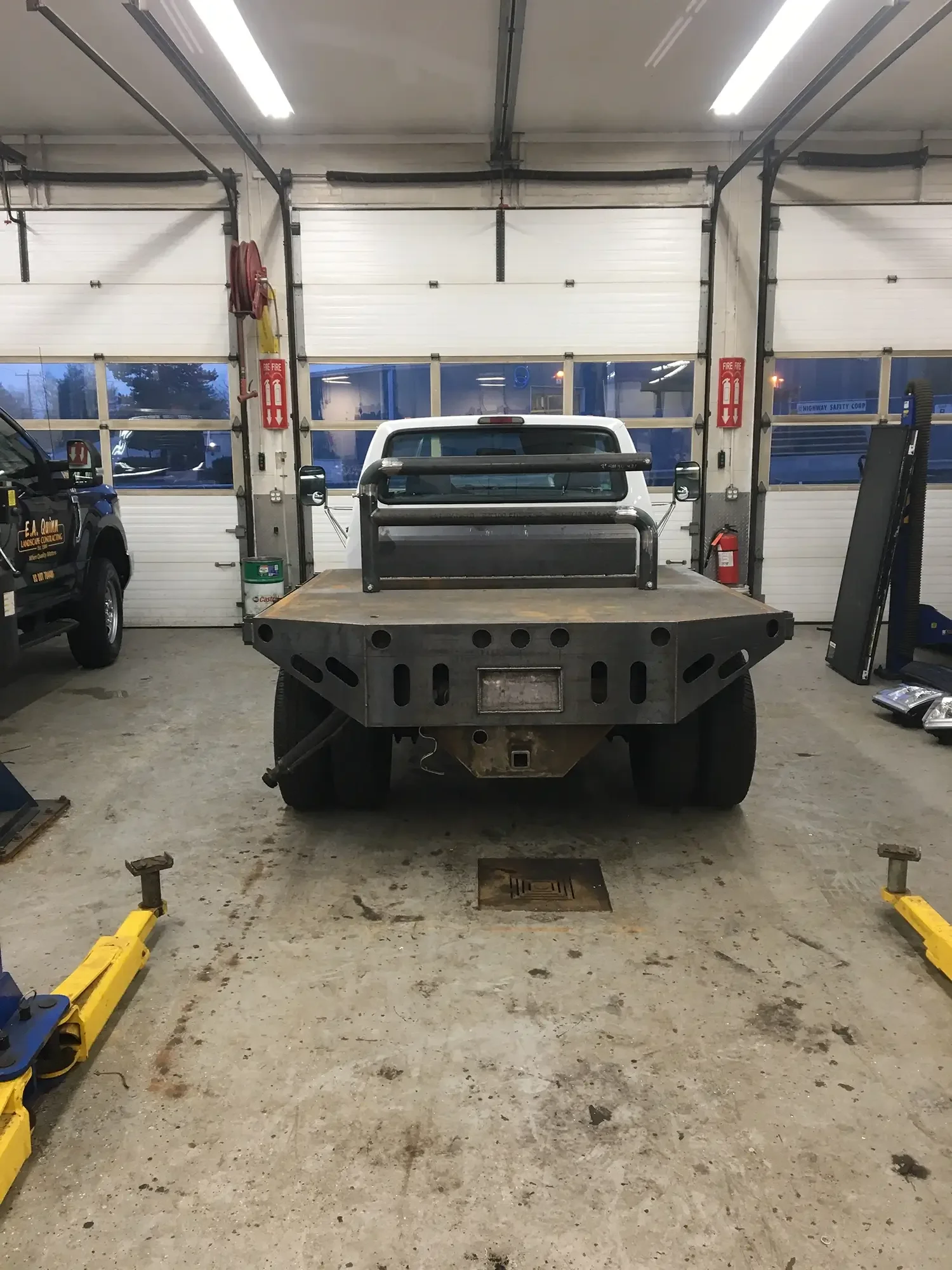 Inside a garage with a white truck on a flatbed, surrounded by tools and equipment, concrete floor with some dirt and stains, garage doors in the background, electrical conduit and signs visible.