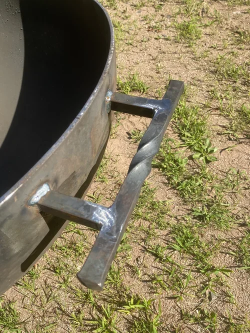 Close-up of a metal trash can with a black handle on a patch of dirt and grass.
