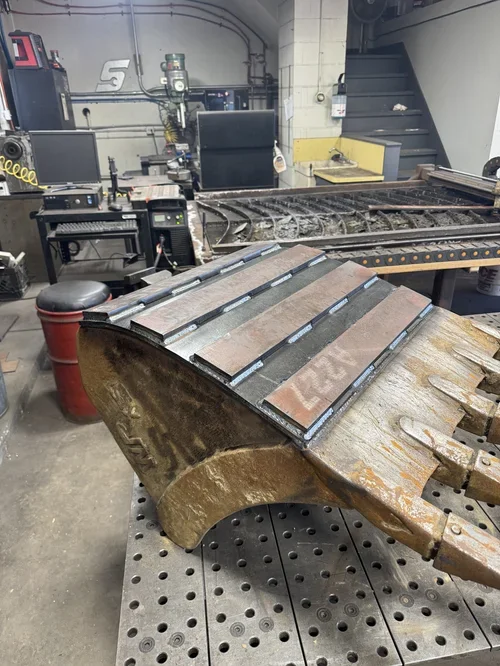 Close-up of a large welding robot's curved metal arm with welding strips, in a workshop with computer monitors, control units, and a track for testing robot movement.