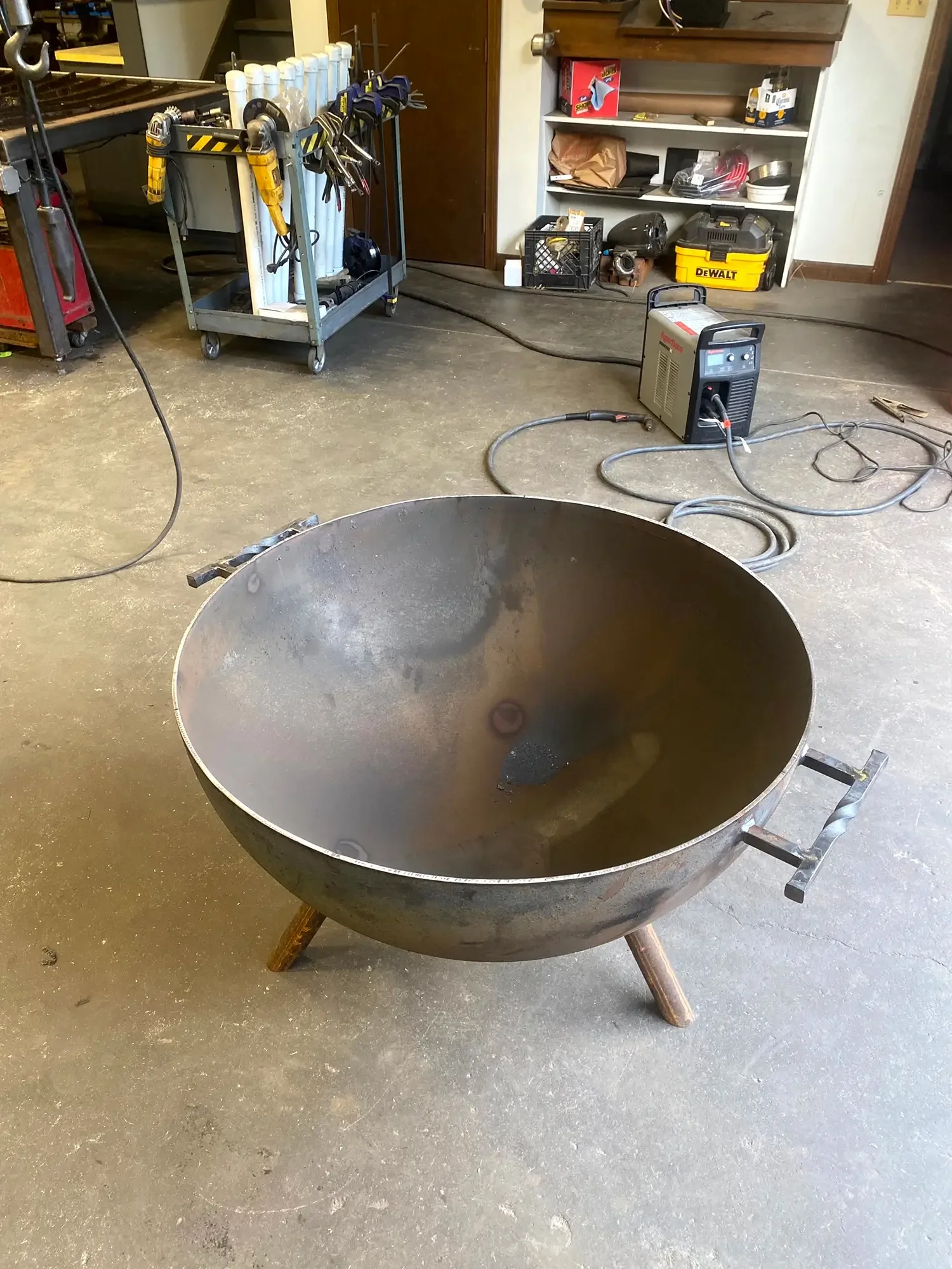 Large metal tub or cauldron with wooden legs, positioned on a concrete floor in a workshop or garage setting.