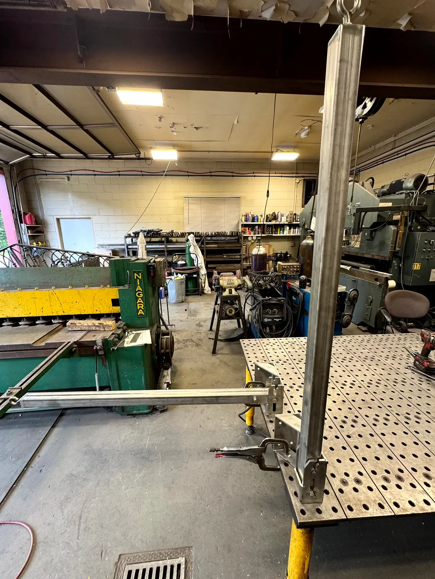 A workshop with various tools and equipment, including a large metal frame in the foreground, a workbench with supplies and bottles on the shelves in the background, and a partially visible garage door on the left side.