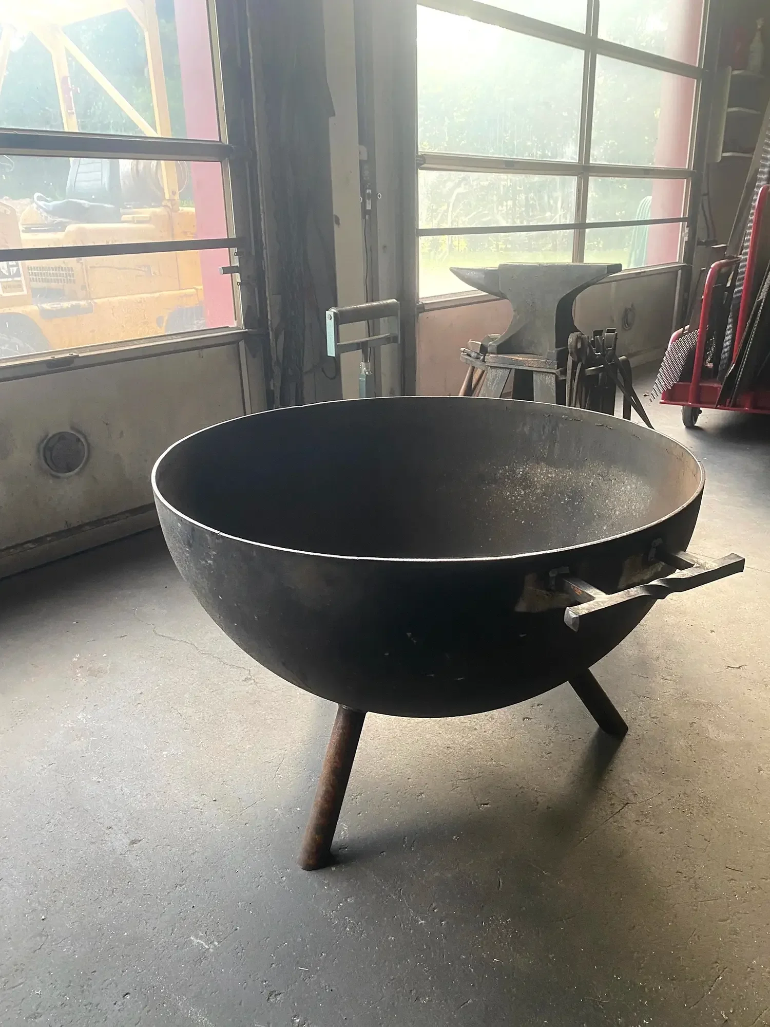 A black metal anvil on a stand in a workshop, with a large metal bowl in the foreground and large windows letting in natural light.