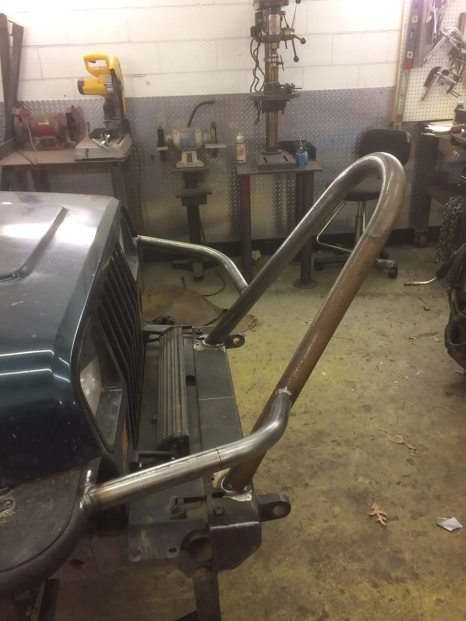 Garage workshop with several tools, including a drill press, grinder, and various hand tools on pegboards. There is a car front end partially visible in the foreground and a metal tubular frame on a workbench. The floor is concrete with some leaves s