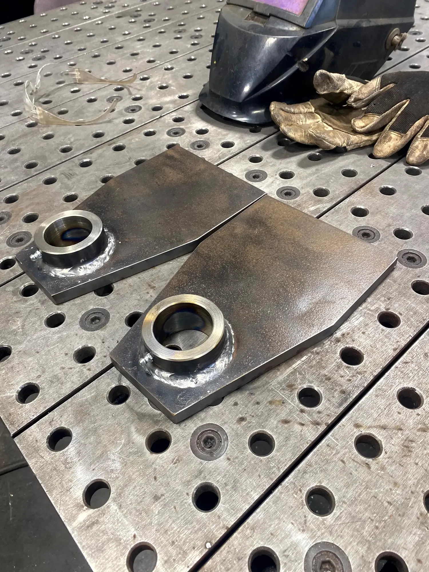 Metal welding work in progress on two rectangular metal pieces with circular holes, placed on a perforated metal work table. Welding gloves, a welding helmet, and safety glasses are nearby.