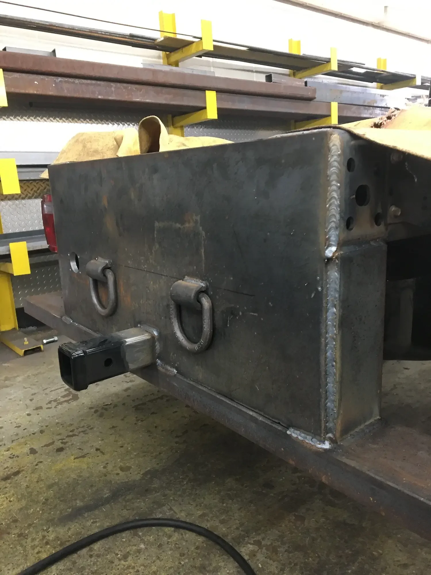 A large metal container with welded seams and attachment points is sitting on a concrete floor, with a yellow strap and a metal shelving unit in the background.