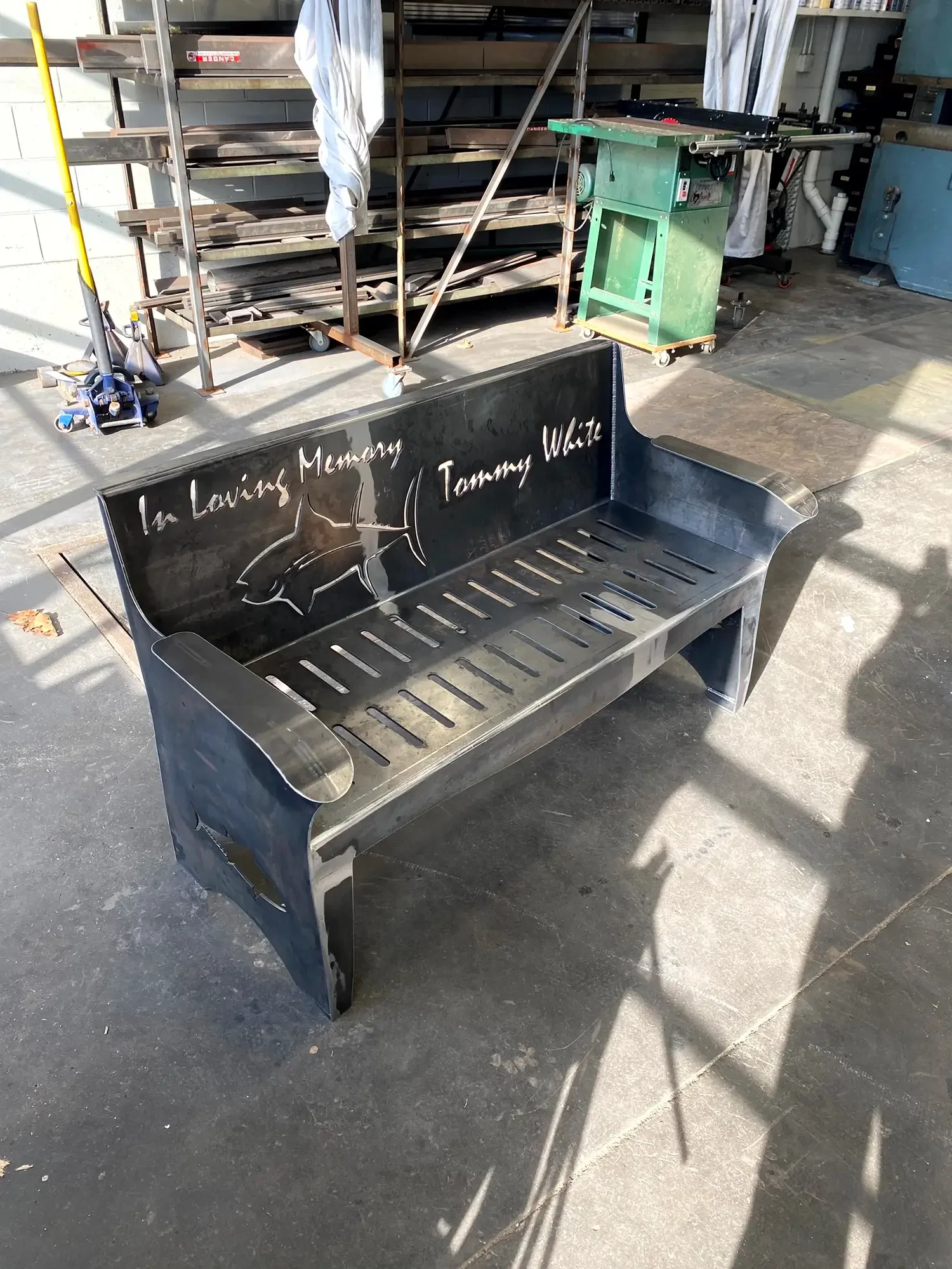 A metal bench with the words 'In Loving Memory Tommy White' engraved on the backrest, placed on a concrete surface outdoors with shadows cast on it.