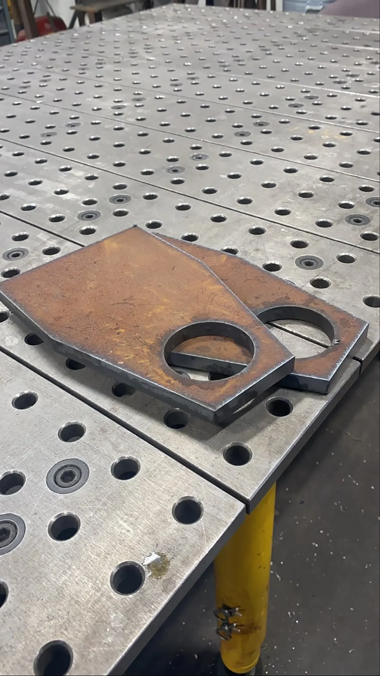 Metal work table with numerous holes and two rusted metal plates with circular cutouts resting on it, supported by a yellow stand.