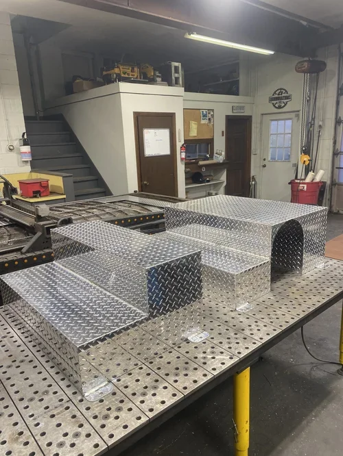 Two custom-made, shiny metal covers with arched openings placed on a perforated workbench inside a workshop or garage.