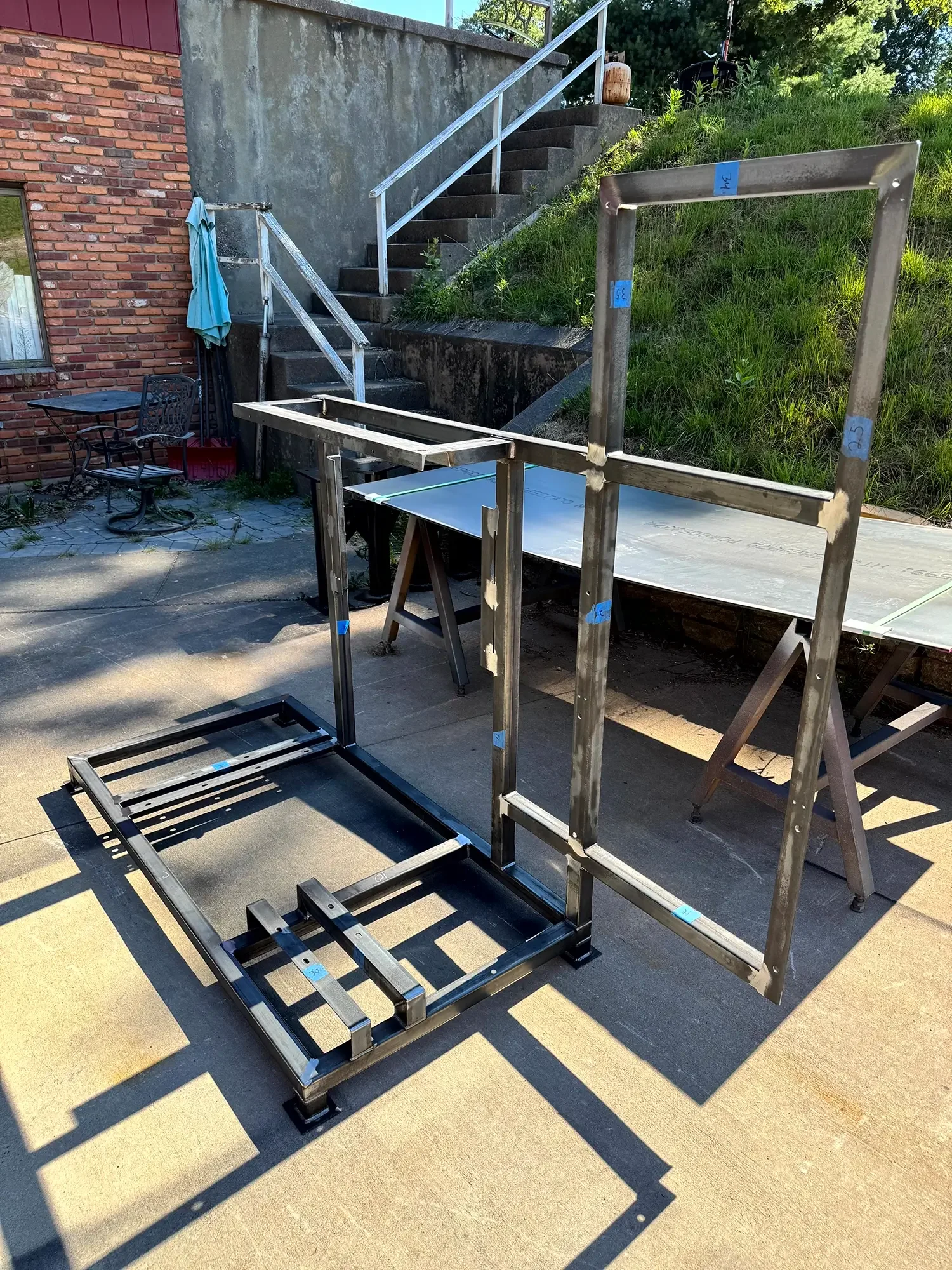 Metal frame structure being assembled outdoors on a paved area, with a brick wall, stairs, green grass, and trees in the background.