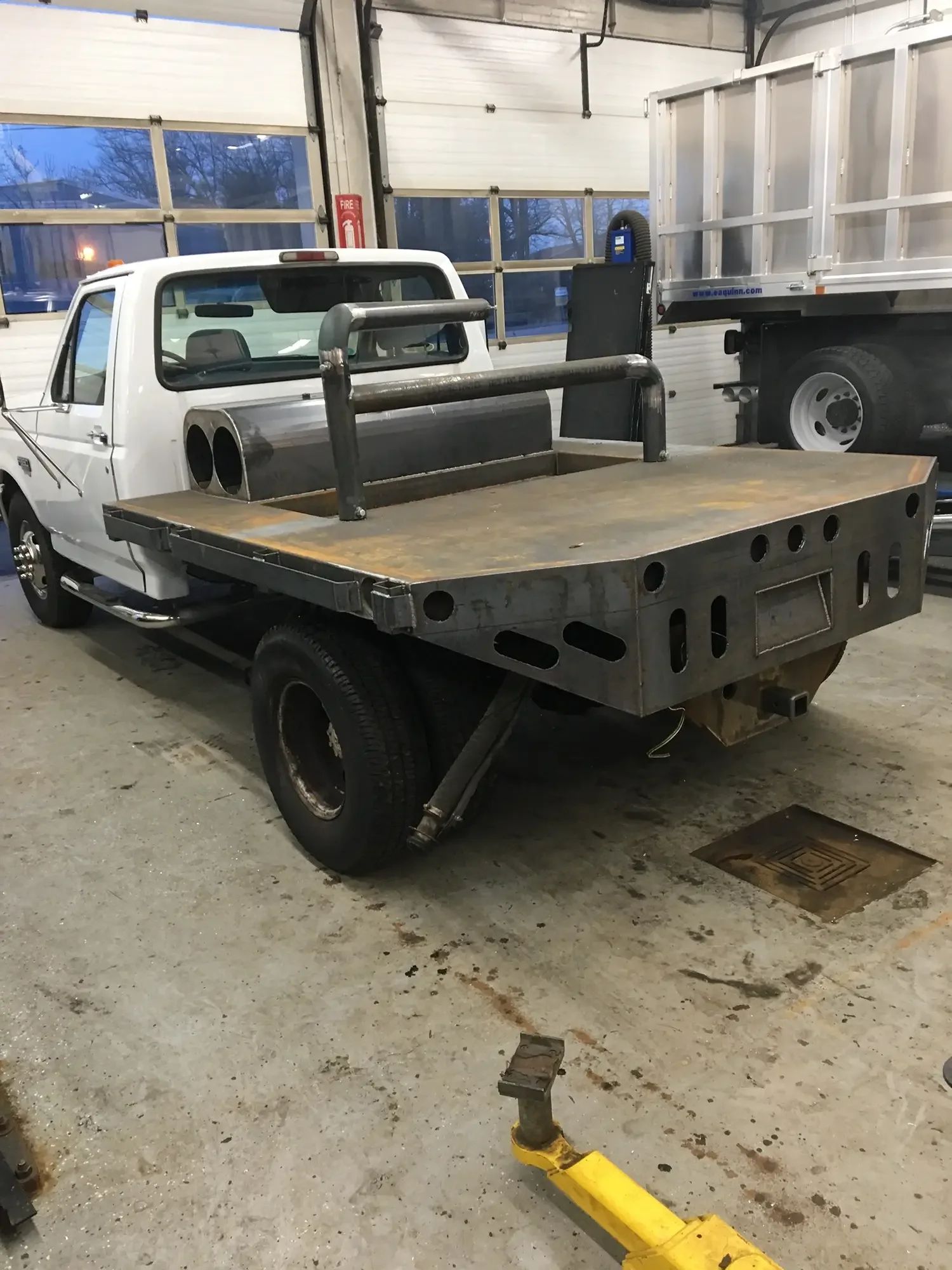 Inside a garage, a small white pickup truck with a flatbed is parked. The flatbed has a metal frame, and there's a large truck with a silver cargo container parked next to it. The garage has large windows with a snowy outdoor scene visible through th