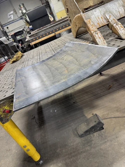 A metal sheet with a curved shape resting on a workbench in a workshop with tools, machinery, and equipment in the background.
