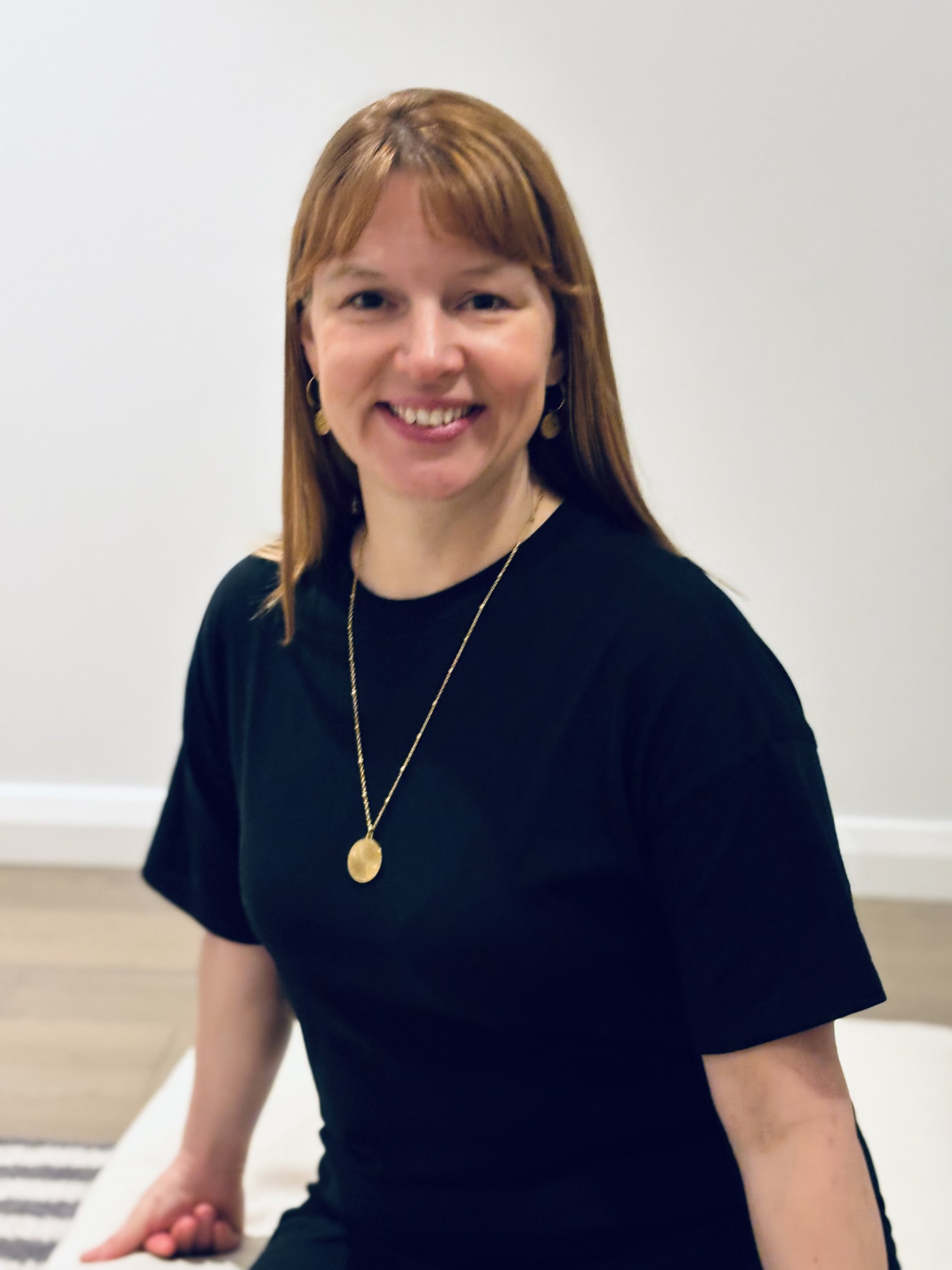 A woman with reddish-brown hair, wearing gold hoop earrings, a black top, and a gold necklace with a circular pendant, sitting against a plain light-colored wall.