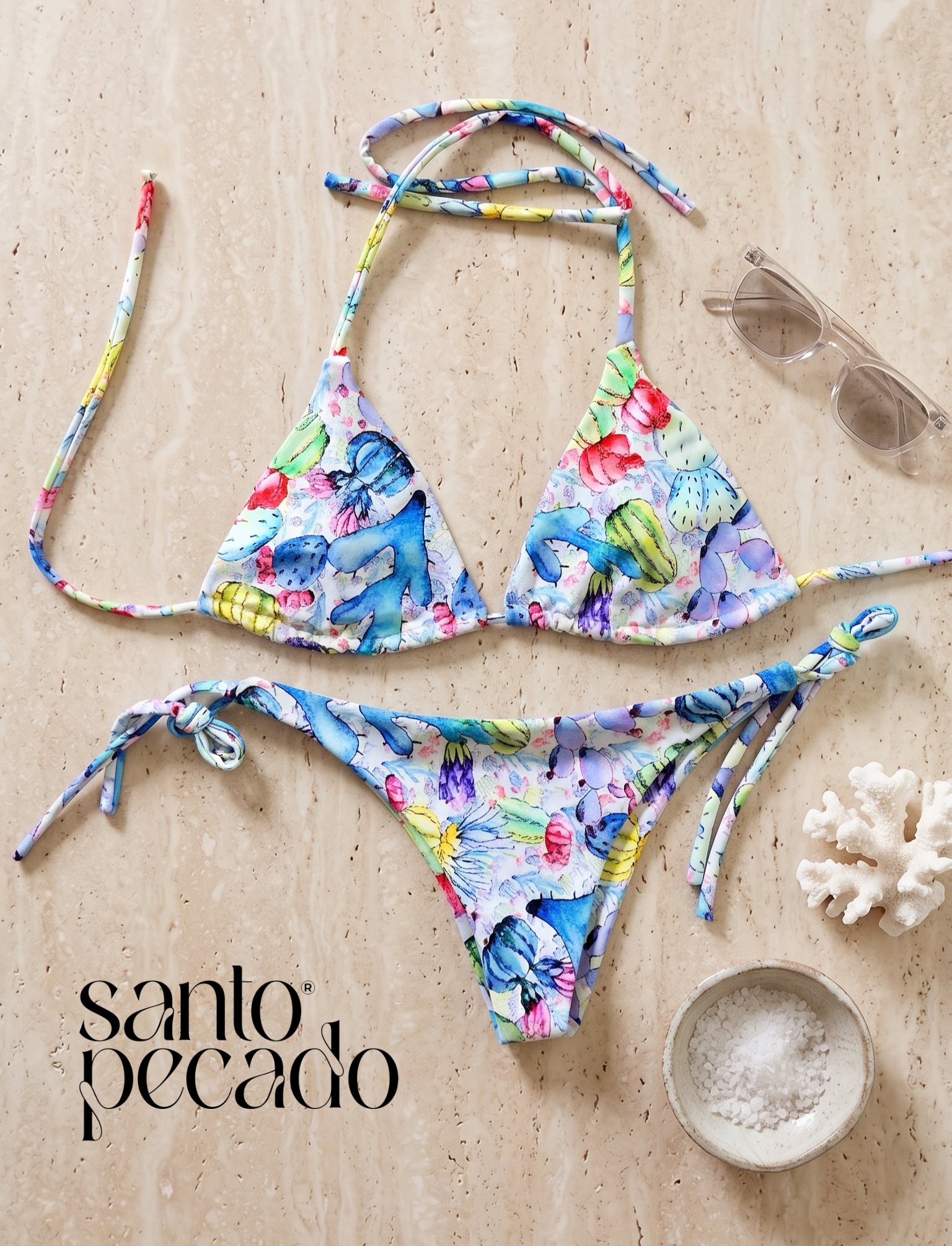 A colorful floral bikini top and bottom set on a beige marble surface, with a small bowl of coarse salt and a white coral decoration nearby.