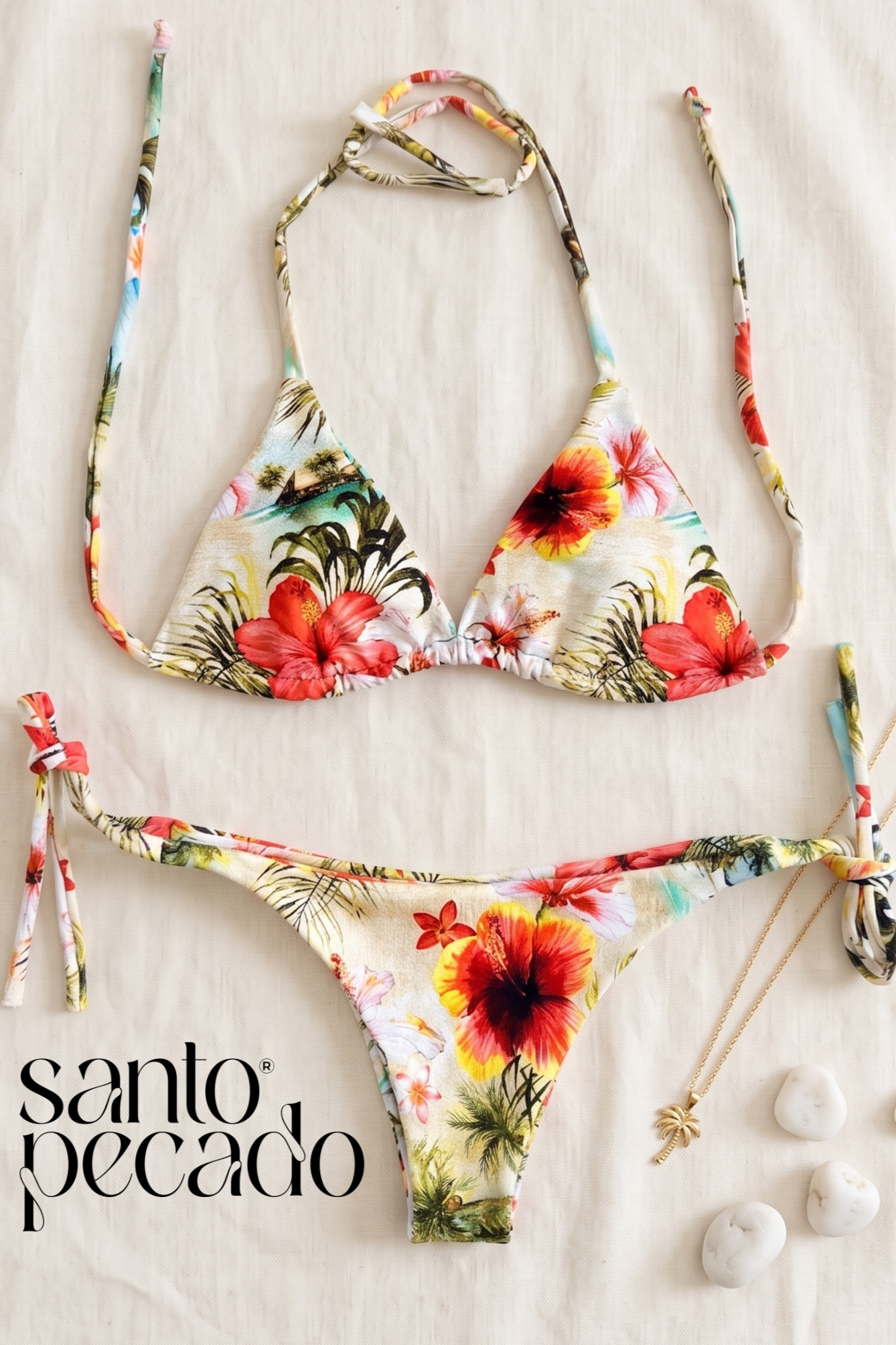 A floral bikini with a tropical island scene and hibiscus flowers on the fabric, laid out on a white surface with a gold palm tree necklace and white stones.