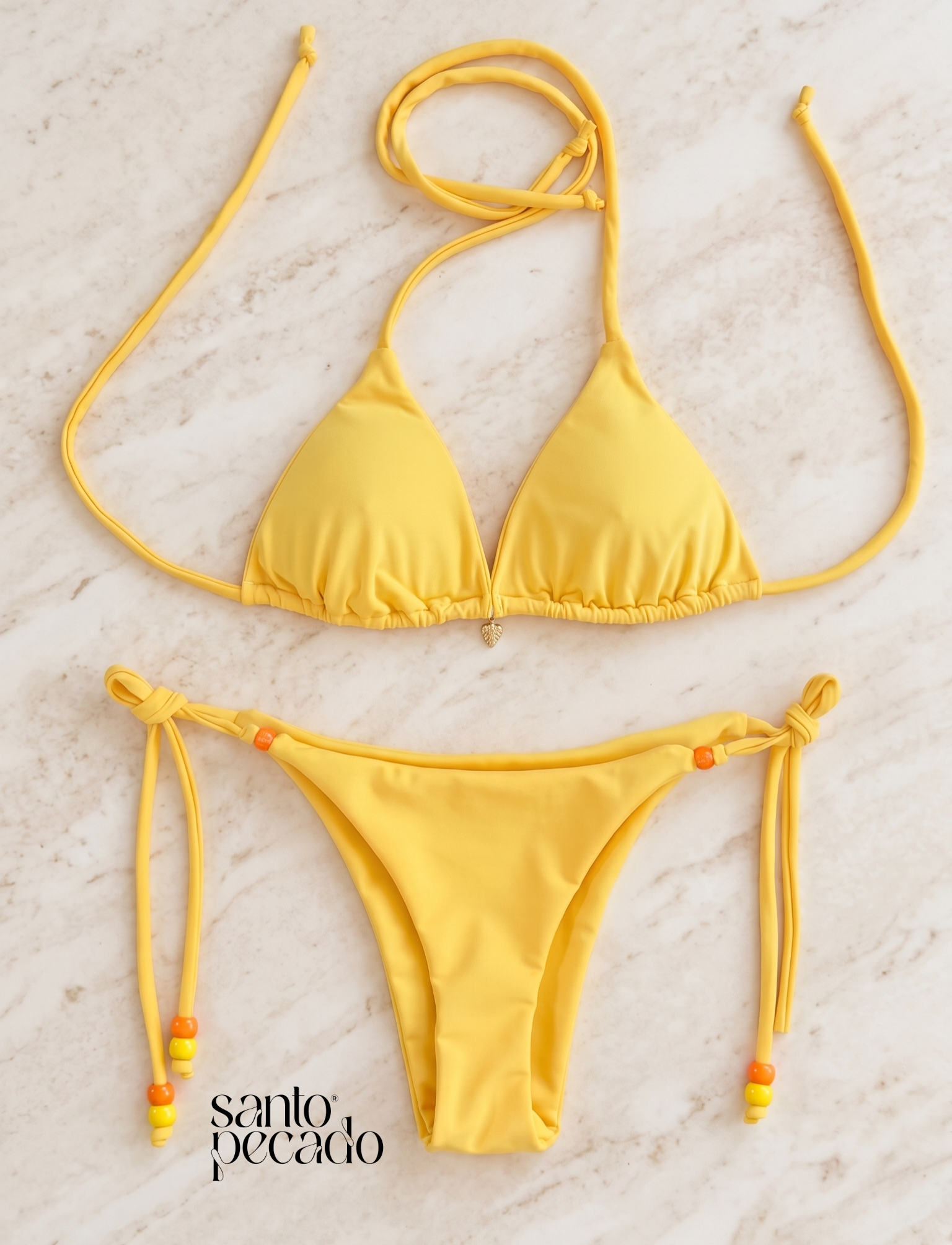 Yellow bikini with string ties and beaded accents laid out on a marble surface, with the words "santo pecado" in the bottom left corner.