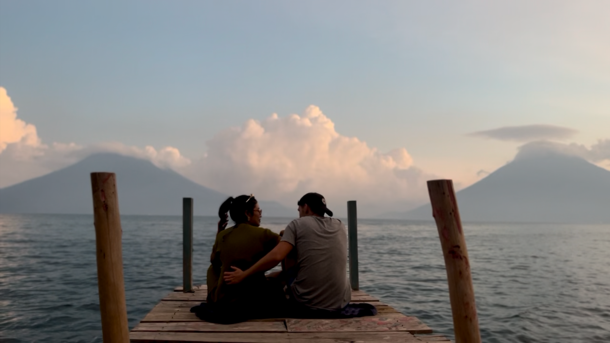 Couple sitting on a wooden dock by the ocean at sunset, with mountains and clouds in the background.