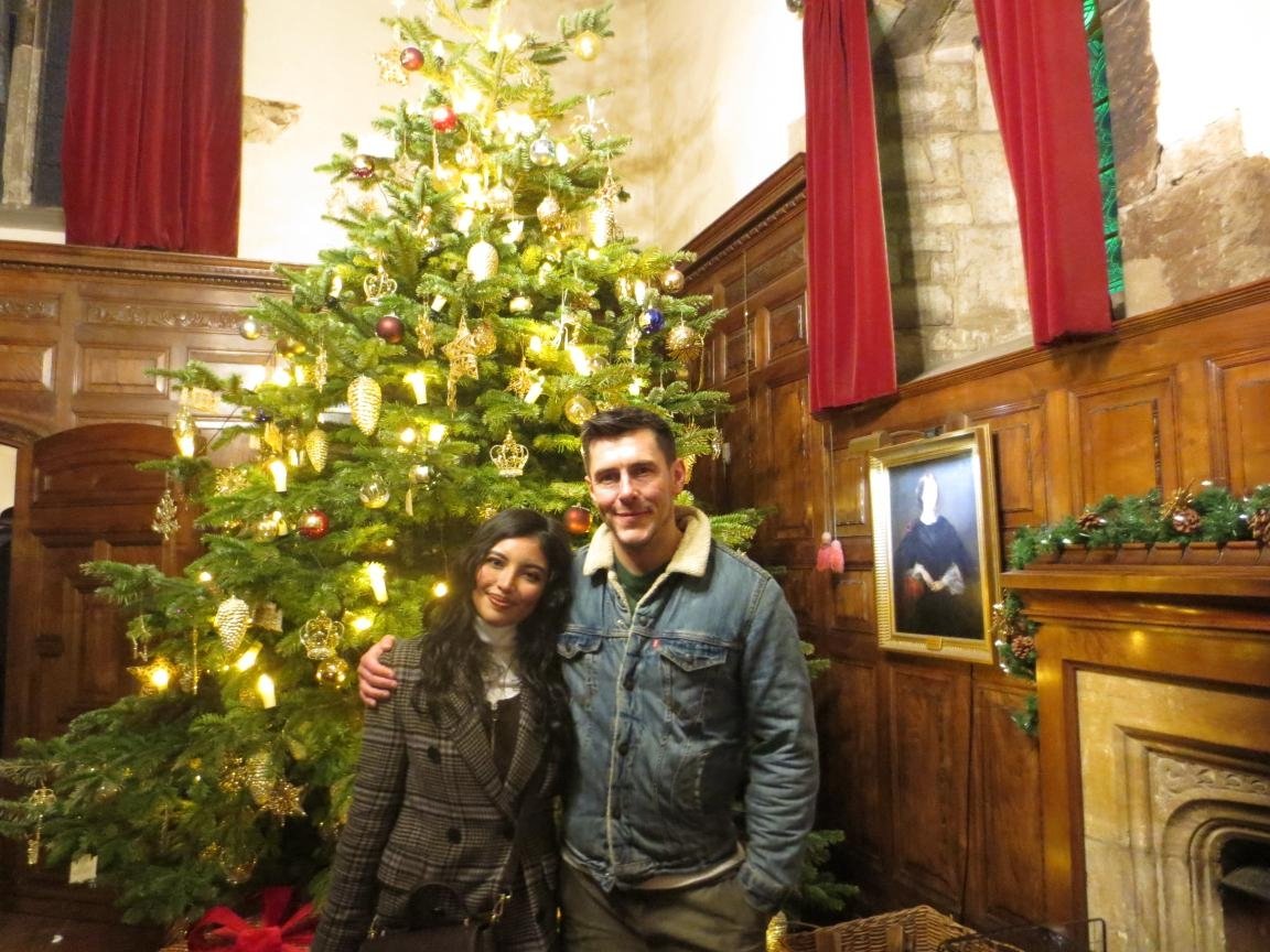 Two people, a woman and a man, standing in front of a decorated Christmas tree in a wood-paneled room with a fireplace, red curtains, and a portrait on the wall.